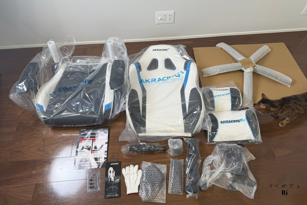 AKRacing Eclairのパーツ一覧。座面・背もたれ・アームレスト・キャスター・ネジ類など、組み立てに必要な部品がすべてそろっている様子。猫も写り込み