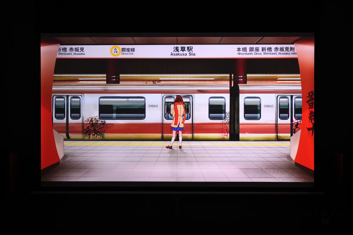 アニメ映像をプロジェクターで投影し、浅草駅のホームに立つキャラクターが鮮やかな色彩で表示されている様子。
