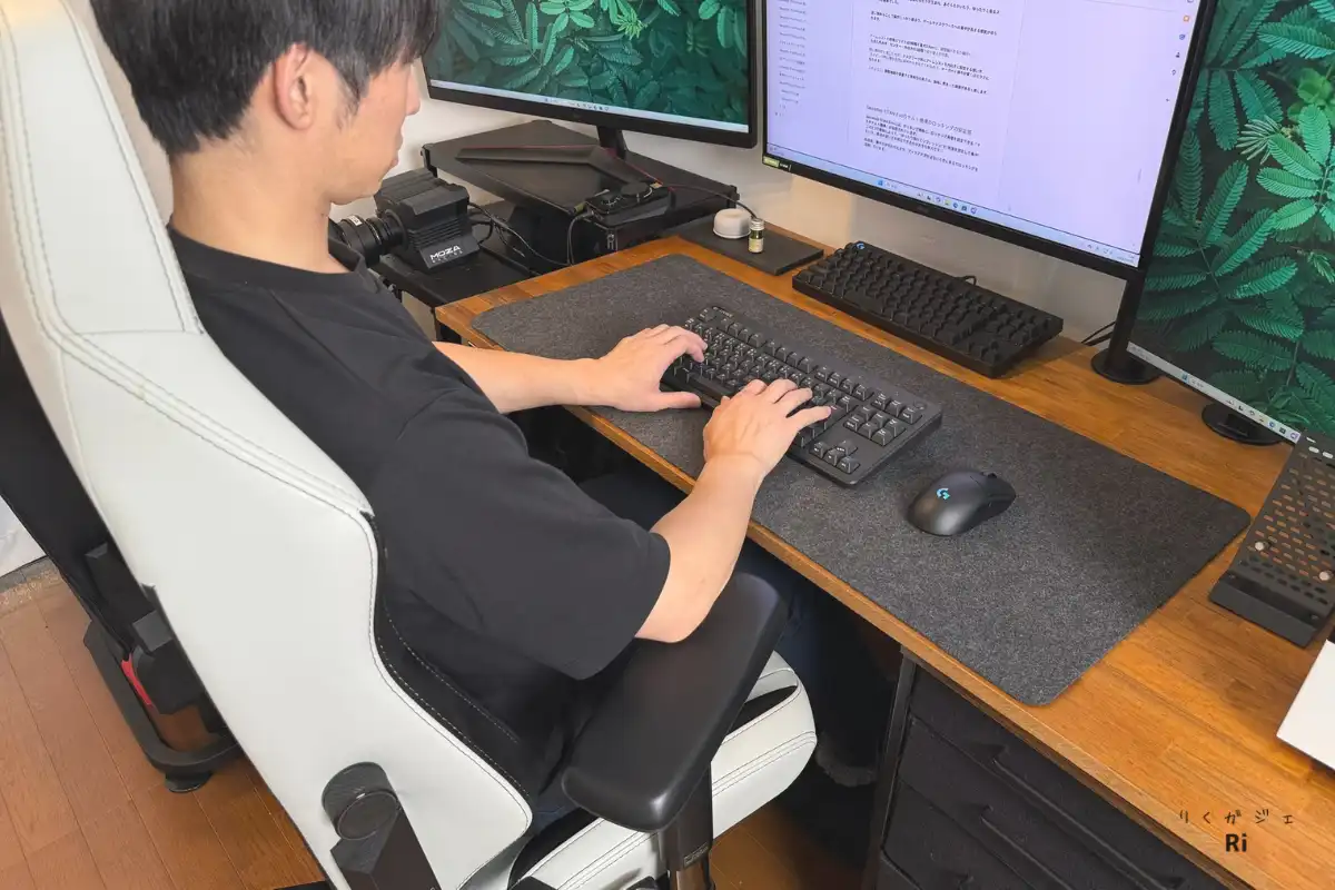 Secretlab TITAN Evoに座り、デュアルモニターの前でタイピングをしている男性。アームレストを内向きに設定し、デスクワークに最適な姿勢で作業している様子。広めのフェルト製デスクマットの上にはキーボードとマウスが配置されている。