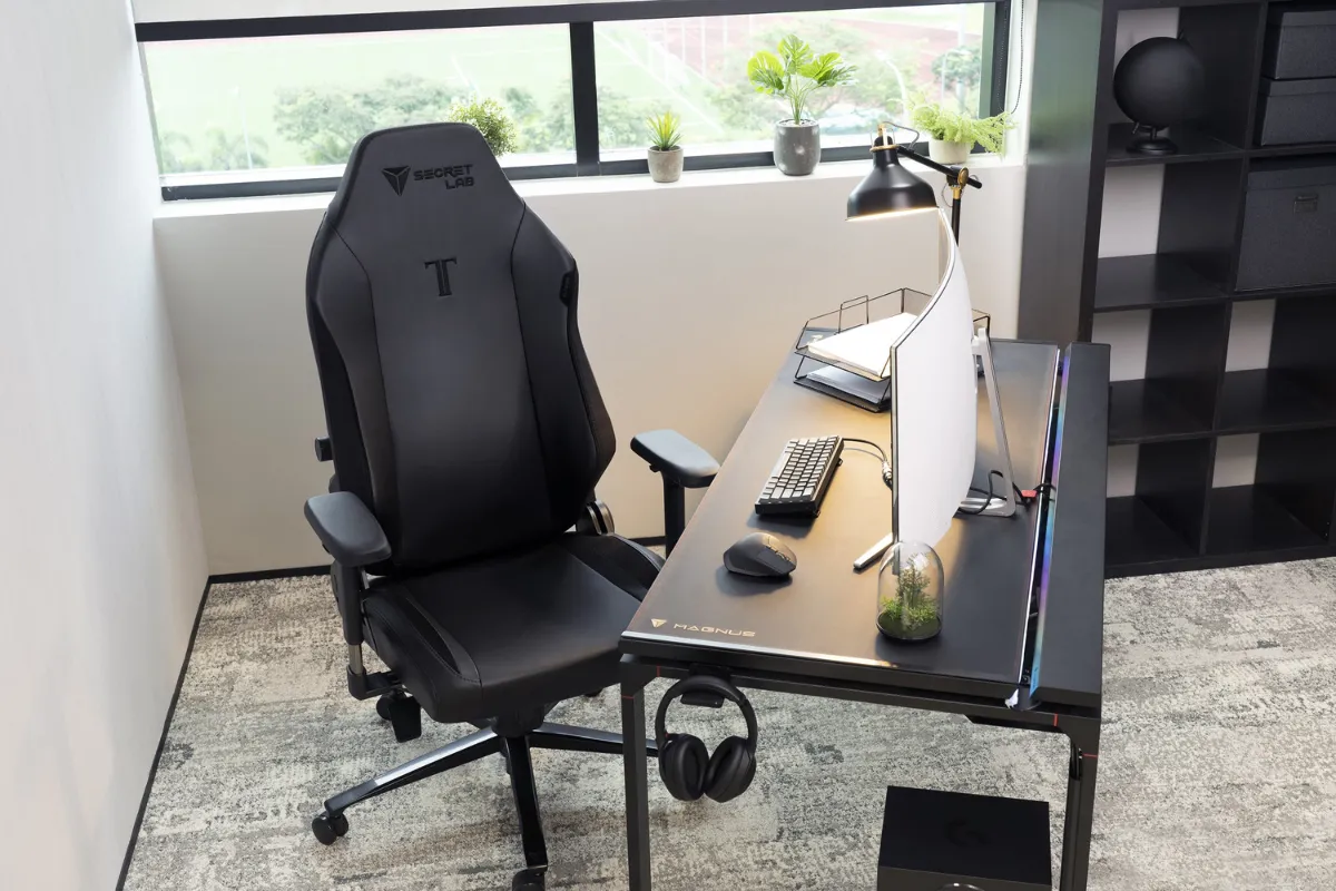 Secretlab TITAN Evoのゲーミングチェアが、シンプルでスタイリッシュなデスク環境に設置されている。ブラックのレザーレット張地のチェアがデスクの前に配置され、デスク上にはモニター、キーボード、観葉植物などが並んでいる。