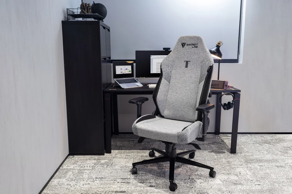 Secretlab TITAN Evoのファブリック張地モデルが、シンプルで落ち着いた雰囲気のワークスペースに設置されている。グレーのチェアが中央に置かれ、背後には黒のデスクとモニター、ノートパソコン、小物が整然と配置されている。