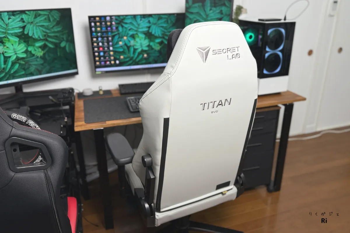 Secretlab TITAN Evoのホワイトモデルを背面から撮影した写真。チェアはL字デスクの前に配置され、3枚のモニターと白いゲーミングPCが設置された環境。左側には別ブランドの黒赤のゲーミングチェアも見える。