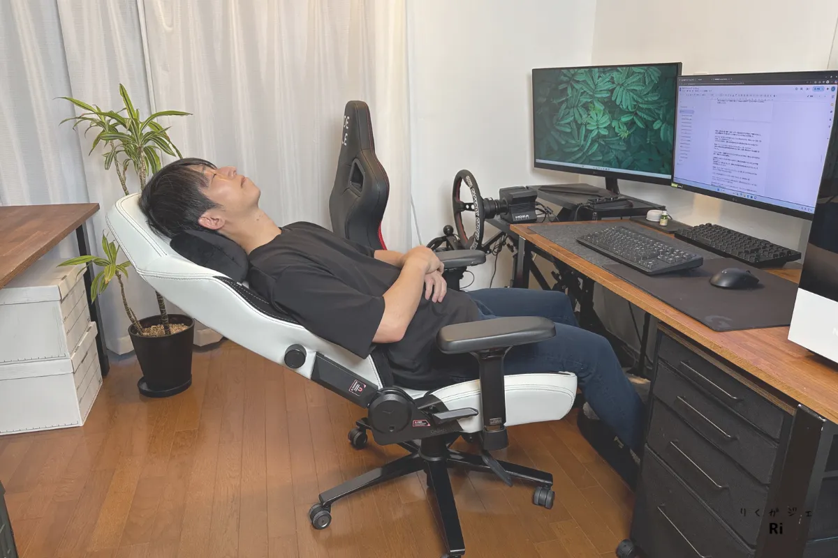 Secretlab TITAN Evoのホワイトモデルで、リクライニングを最大まで倒して仰向けにくつろぐ男性。L字型デスクにはデュアルモニターとキーボードが設置されており、作業とリラックスを両立できるPC環境になっている。