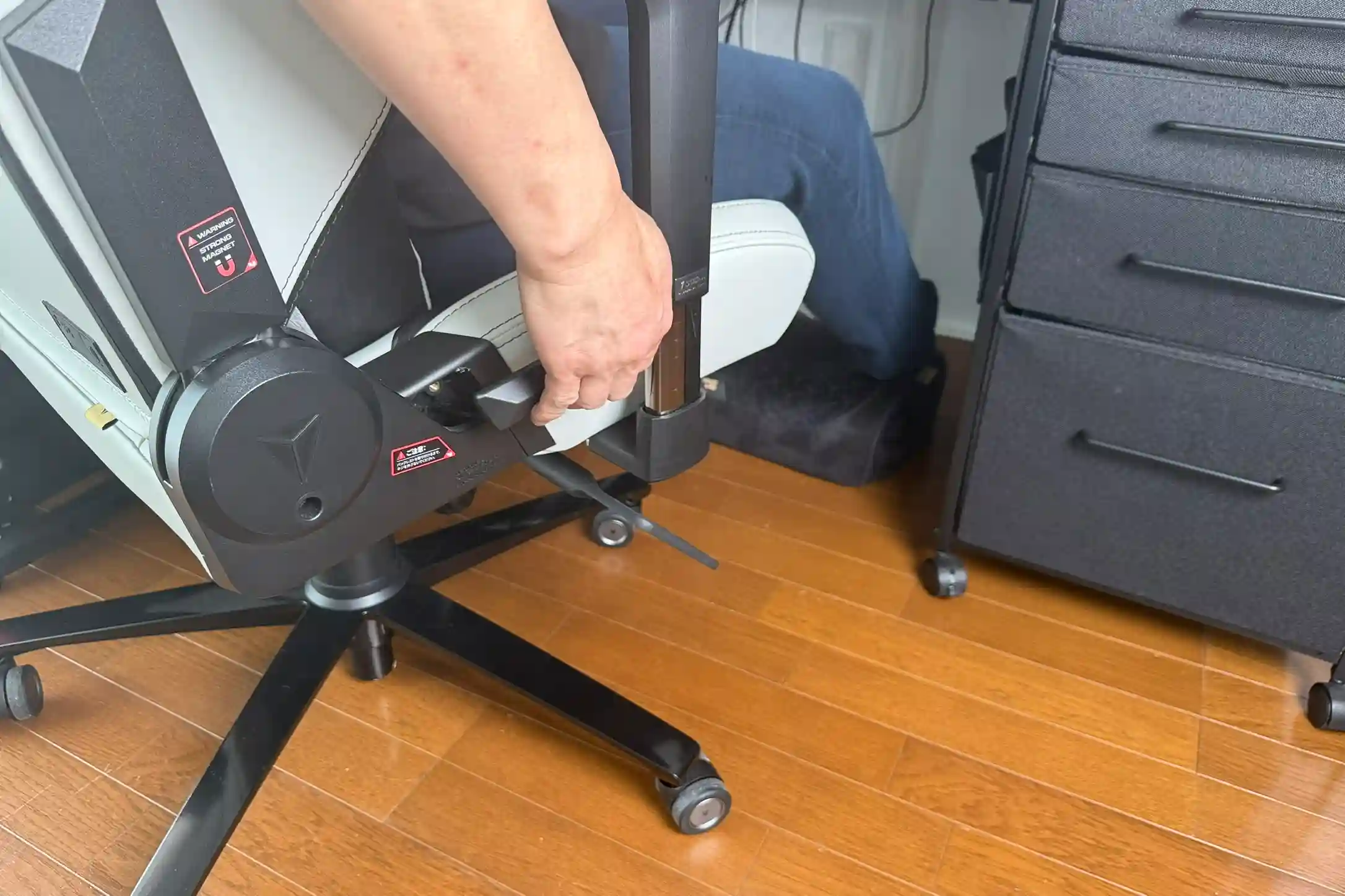Secretlab TITAN Evoのゲーミングチェアで、座面左下にあるリクライニングレバーを操作している様子。チェアに座った状態でリクライニング角度を調整している場面が、床とデスク周辺を含めて映されている。